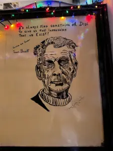 A black-and-white portrait drawing of a man with strong features is on a wall in a pub, alongside a quote from Samuel Beckett and a Guinness pint glass under colorful string lights. The quote reads, “We always find something, ah Didi, to give us the impression that we exist?”.