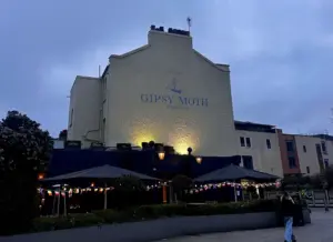 A yellow-painted building at dusk displays "Gipsy Moth Greenwich" with a ship logo. Outdoor seating under umbrellas and string lights is in front; a person walks nearby. Bushes and modern buildings are in the background.