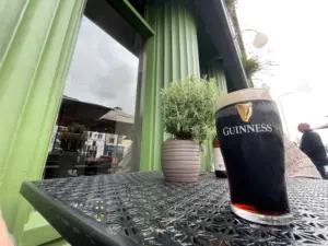 A Guinness pint glass sits on a black metal outdoor table near a potted plant outside a green pub, with reflections in the window and people in the background.
