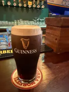 A Guinness pint glass with a creamy head sits on a beer mat on a pub counter, with glasses and bar taps visible in the background.