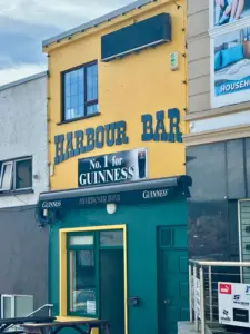 A yellow and green building with a sign reading "HARBOUR BAR" features a Guinness pint glass illustration above the entrance. Surrounded by other buildings, it has a small outdoor seating area.