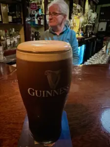 A Guinness pint glass sits on a bar counter, with a woman in a blue top standing behind the pub bar surrounded by bottles and glasses.
