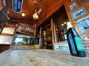 A rustic, wooden-walled room decorated with nautical items and boat models. A hand holds a Guinness pint glass on a weathered wooden table, with framed pictures, signs, and warm lighting completing the cozy pub vibe.