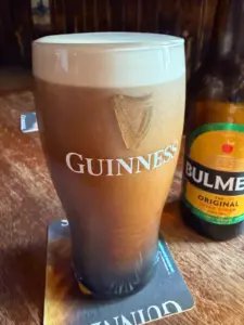 A pint glass of Guinness stout with a creamy head sits on a coaster next to a bottle of Bulmers Irish cider on a wooden table in a cozy pub setting.