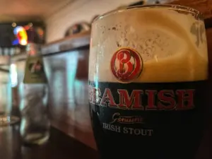 A close-up of a pint glass filled with Beamish Genuine Irish Stout, showing a creamy head. In the background, there is a blurred bottle and bar counter, giving a cozy pub atmosphere.
