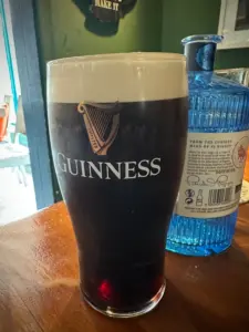A pint glass of Guinness stout with a creamy head sits on a wooden table next to a textured blue gin bottle in a cozy, well-lit bar setting.