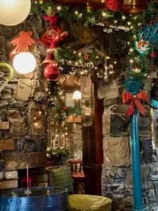 A cozy stone-walled room decorated with Christmas garlands, red bows, ornaments, and a Guinness pint glass. Warm lights illuminate a doorway labeled "PRESIDENT'S SNUG," leading to a festive, inviting pub interior.