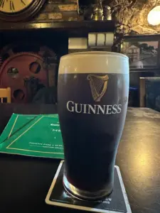 A Guinness pint glass on a coaster rests on a dark table in a cozy pub, with a green menu nearby and rustic decor featuring red gears and a large clock in the background.