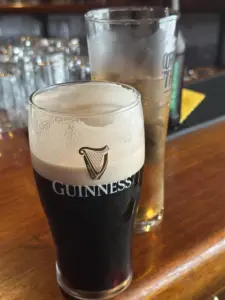 A Guinness pint glass with a creamy head sits on a wooden bar beside a tall glass with ice, both in front of glassware and pub equipment.