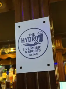 A sign on a wooden pillar reads "The Hydro" with a Guinness pint glass icon, and "Live Music & Sports Est. 2025" in bold letters. The background shows the cozy interior of a pub with bottles visible.