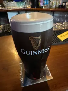 A Guinness pint glass with a creamy head rests on a Guinness-branded coaster at the pub, with glasses and bottles softly blurred in the background.