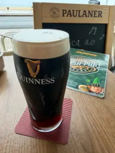 A pint of Guinness stout sits on a red-striped coaster on a wooden table, with an Irish pub menu and a Paulaner sign in the background.