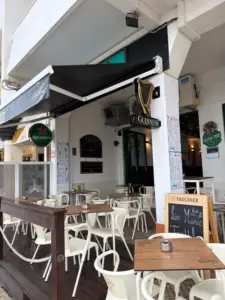 Outdoor seating area of a pub with white chairs and tables, covered by black awnings displaying Heineken and Guinness logos. A chalkboard advertises "Live Music." The pub has white walls and various posters.