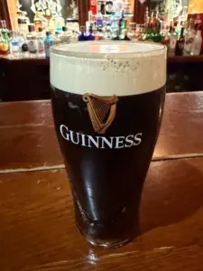 A pint of Guinness stout with a creamy head sits on a wooden bar counter, with bottles of various alcohols blurred in the background.