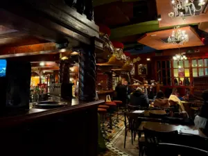 Dimly lit pub with wooden décor, ornate pillars, chandeliers, and groups of people sitting at tables and the bar. Some patrons are engaged in conversation, creating a cozy, lively atmosphere.
