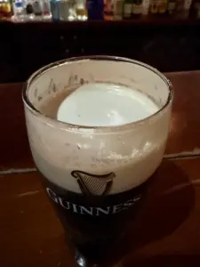 A pint glass of Guinness stout beer with a creamy frothy head sits on a wooden bar, with bottles of liquor visible blurred in the background.