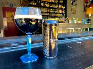 A Guinness pint glass with a creamy head sits beside a tall can of Guinness on a bar counter, with shelves of bottles and barstools visible in the background, evoking the atmosphere of a cozy pub.