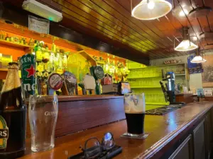 A cozy pub interior with a wooden bar, beer taps, bottled drinks, and glasses. A pint of dark beer and an empty glass sit on the bar, near a pair of eyeglasses. Warm lighting creates a welcoming atmosphere.