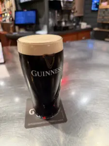A pint of Guinness stout with a creamy head sits on a coaster atop a shiny metal bar counter, with blurred bar equipment and screens in the background.