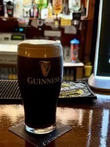 A Guinness pint glass with a thick, creamy head sits on a coaster at the pub’s wooden bar, with bottles and bar equipment softly blurred in the background.