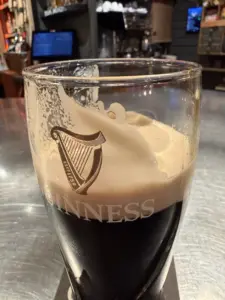 A partially drunk pint of Guinness beer with a creamy foam head sits on a bar counter, with the Guinness harp logo visible on the glass and bar shelves blurred in the background.