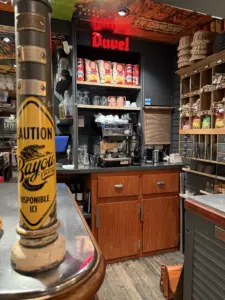 A cozy bar counter with a yellow “Caution Bayou” beer tap in the foreground, a coffee machine, shelves with cups, Duvel neon sign, and wooden shelves with food items in the background.