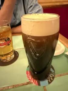 A pint of dark beer with a creamy head sits on a colorful coaster next to a half-full glass of light beer on a tiled table; a person with crossed arms is partially visible in the background.