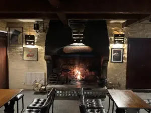 A cozy pub interior with wooden tables and chairs, a large stone fireplace with a fire burning, and warm lighting on textured walls decorated with framed pictures and rustic decor.