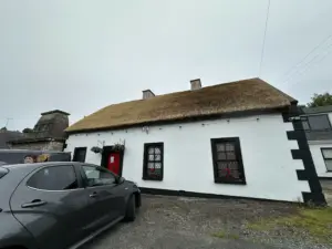 A white cottage with a thatched roof, black-framed windows, and a red door. A grey car is parked in front, and the sky is overcast. Two people are partly visible near the car.