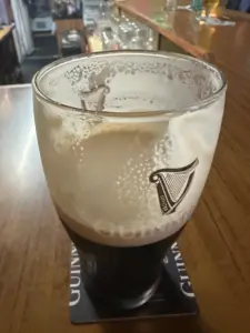 A partially drunk pint of Guinness sits on a branded coaster atop a wooden bar, with foam clinging to the inside of the glass. The bar and glassware are visible in the background.