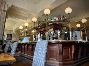 A cozy pub interior with a wooden bar, globe light fixtures, and shelves stocked with bottles. Plaid-upholstered chairs and tables are in the foreground, and soft lighting creates a warm atmosphere.