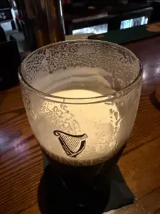 A partially finished Guinness pint glass sits on a black napkin on a wooden bar, with foam lacing inside the glass and pub shelves in the background.