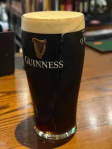 A Guinness pint glass with a creamy head sits on a wooden bar counter in a cozy pub, with bar equipment and a blurred background visible.