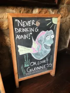 A chalkboard sign shows a cheerful fish with “Never drinking again!” above and “Oh, look! Guinness” below, referencing Dory from Finding Nemo. A Guinness pint glass appears next to sun and seaweed, adding to the pub charm.