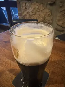 A Guinness pint glass with a thick creamy head sits on a wooden table in a cozy pub, with a stone wall and window in the background.