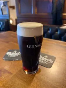 A guinness pint glass with a creamy head sits on a wooden pub table, Carlsberg-branded coasters nearby and dark wooden furniture in the background.