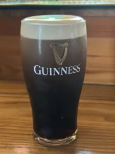 A Guinness pint glass with a creamy head sits on a wooden pub counter. The Guinness logo and harp are visible on the glass, with a softly blurred background.