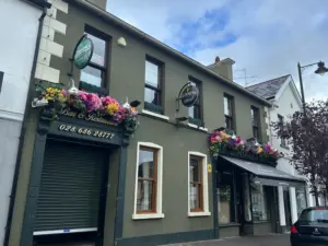A two-story olive green building with flower boxes on the upper windows, a closed shutter on the left, and signs for a pub and restaurant. A Guinness pint glass is displayed outside. The sky is partly cloudy and a tree stands to the right.