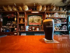 A Guinness pint glass sits on a wooden bar counter in a cozy, traditional Irish pub, with shelves of bottles, mugs, and jugs in the background and a vintage sign on the wall.