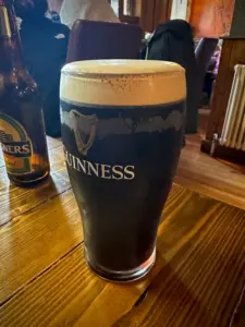 A Guinness pint glass with a creamy head sits on a wooden table in a cozy pub, with a bottle of Magners cider and people seated in the background.