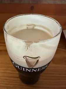 A partially finished Guinness pint glass sits on a wooden table in a cozy pub, with foam residue clinging to the inside and the iconic Guinness logo visible on the front.