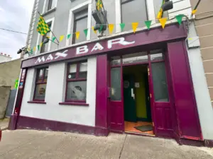 Street view of "Max Bar," a pub with maroon trim, white walls, and green and yellow flag bunting. A Guinness pint glass logo is visible by the open entrance, offering a glimpse of the pub's green-walled interior.