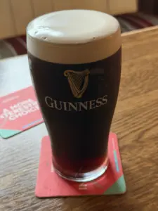 A Guinness pint glass with a creamy head sits on a red coaster on a wooden table, with a blurred red and white coaster in the background, evoking the warm atmosphere of a pub.