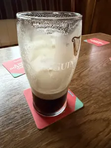 A half-empty Guinness pint glass with foam residue sits on a red coaster on a wooden table in a pub, with more coasters and upholstered seating visible in the background.