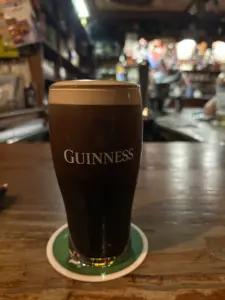 A Guinness pint glass sits on a coaster on a wooden bar counter in a dimly lit pub, with shelves and blurred patrons in the background.