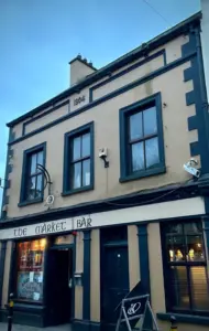 A two-story tan building with dark trim, labeled “1904” and “The Market Bar.” The entrance is below two large windows, and a signboard featuring a Guinness pint glass stands outside. A window displays posters inside the pub.