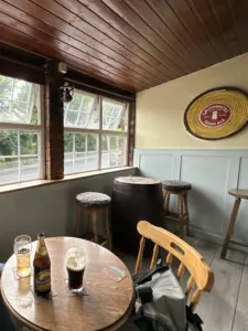 A cozy pub corner with a wooden ceiling, a round table holding a Guinness pint glass, a backpack on a chair, barrel-shaped table with stools, large windows, and a Smithwick's Irish Ale sign on the wall.