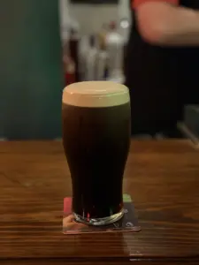 A Guinness pint glass with a creamy head sits on a coaster atop a wooden pub bar, with a blurred background and dim lighting.