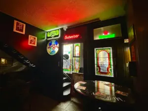 Dimly lit pub interior with neon Carlsberg and Budweiser signs above a door. Stained glass panels feature colorful designs. Photos hang on the dark wall, and a round table with papers and a Guinness pint glass sits in the foreground.