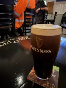 A Guinness pint glass rests on a coaster atop a dark bar table in a pub, with people seated in the background and part of a sign reading "BAR" visible on the table.